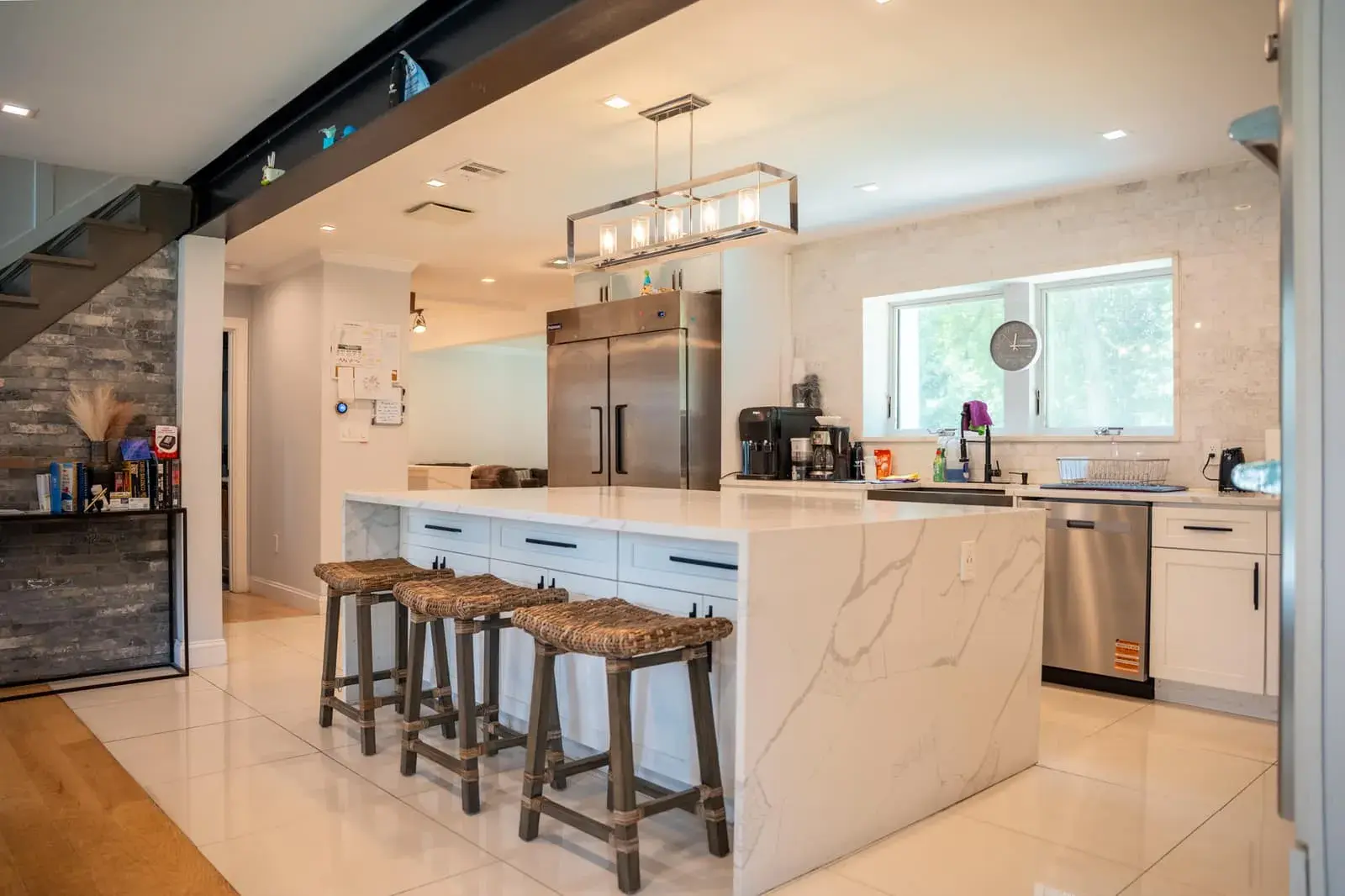 The kitchen at Closter Residence I: a long marble island with woven stools, white cabinets and a steel refrigerator